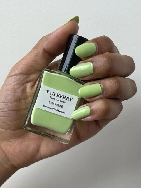 Eine Hand mit grün bestrichenen Nageln. Der Nagellack dazu hält sie so in der Hand, damit ihre Nägel gut sichtbar sind. Die Farbe vom Nagellack ist Grün und auf der Flasche hat es ein Etikett mit dem Namen 