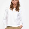 Modell trägt eine klassische weisse Bluse, zusammen mit einer hellbraunen Hose. Ihre Hände sind in den Seitentaschen. Sie trägt die Haar offen und hat sie hinter ihren Ohren gesteckt. Ihre langen Ohrringe kommen in den Haaren hervor. Sie hat den Mund leicht geöffnet und zeigt einen gleichgültigen Ausdruck.