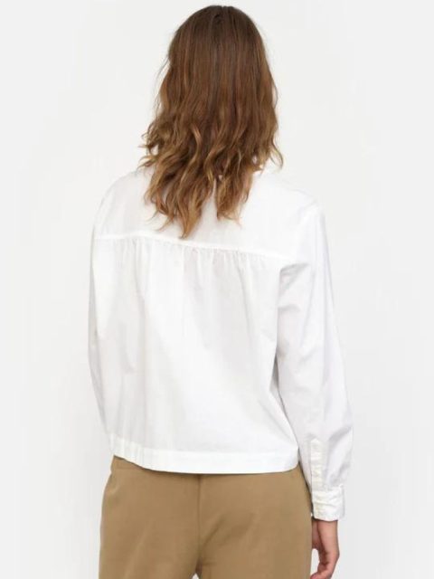 Modell ist von hinten ersichtlich. Sie trägt eine klassische weisse Bluse mit hellbraunen Hosen und hat offenes braunes Haar