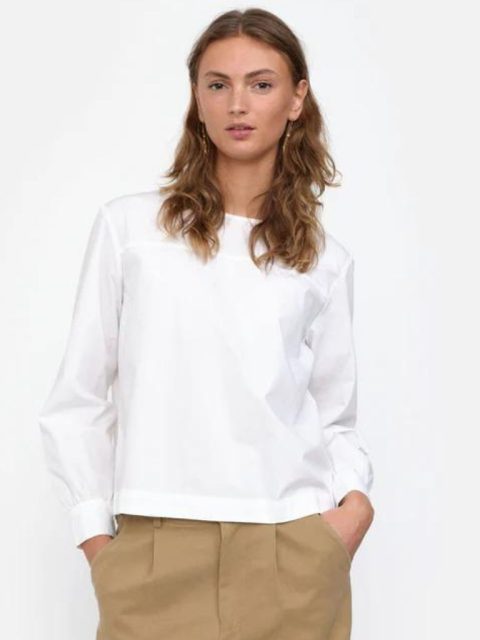 Modell trägt eine klassische weisse Bluse, zusammen mit einer hellbraunen Hose. Ihre Hände sind in den Seitentaschen. Sie trägt die Haar offen und hat sie hinter ihren Ohren gesteckt. Ihre langen Ohrringe kommen in den Haaren hervor. Sie hat den Mund leicht geöffnet und zeigt einen gleichgültigen Ausdruck.
