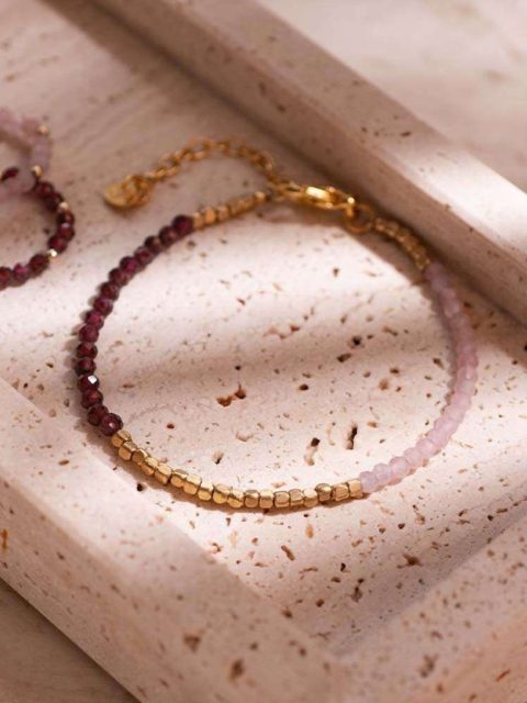 Armband mit kleinen Rosa, Rot und Gold Edelsteinen liegen auf einem unebenen Grund. Das Armband hat einen Verschluss in Gold