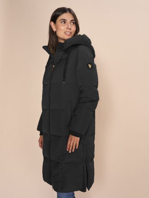 Modell steht seitlich und trägt einen Daunenmantel in Schwarz. Die Jacke hat am linken armen einen kleinen Patch angemacht mit einem Goldherz.