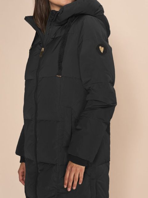 Schwarzer Daunenmantel von der Marke Mos Mosh. Auf dem Bild sieht man nur von der Schulter abwärts ein Modell, dessen Hände sichtbar sind. Am Arm hat es ein Stoffpatch mit einem goldigen Herz.
